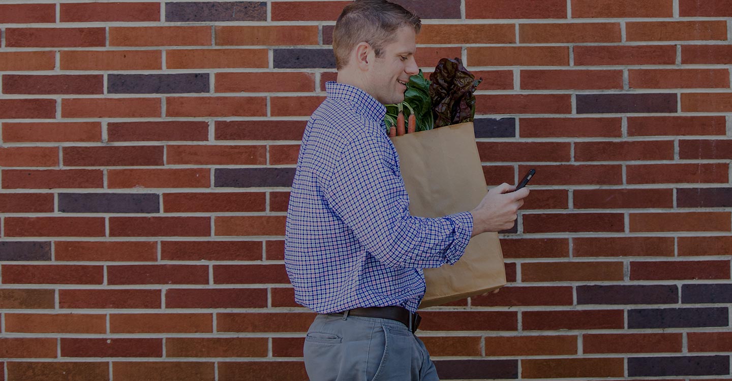 A man looks at his cell phone while carrying a grocery bag full of healthy food options